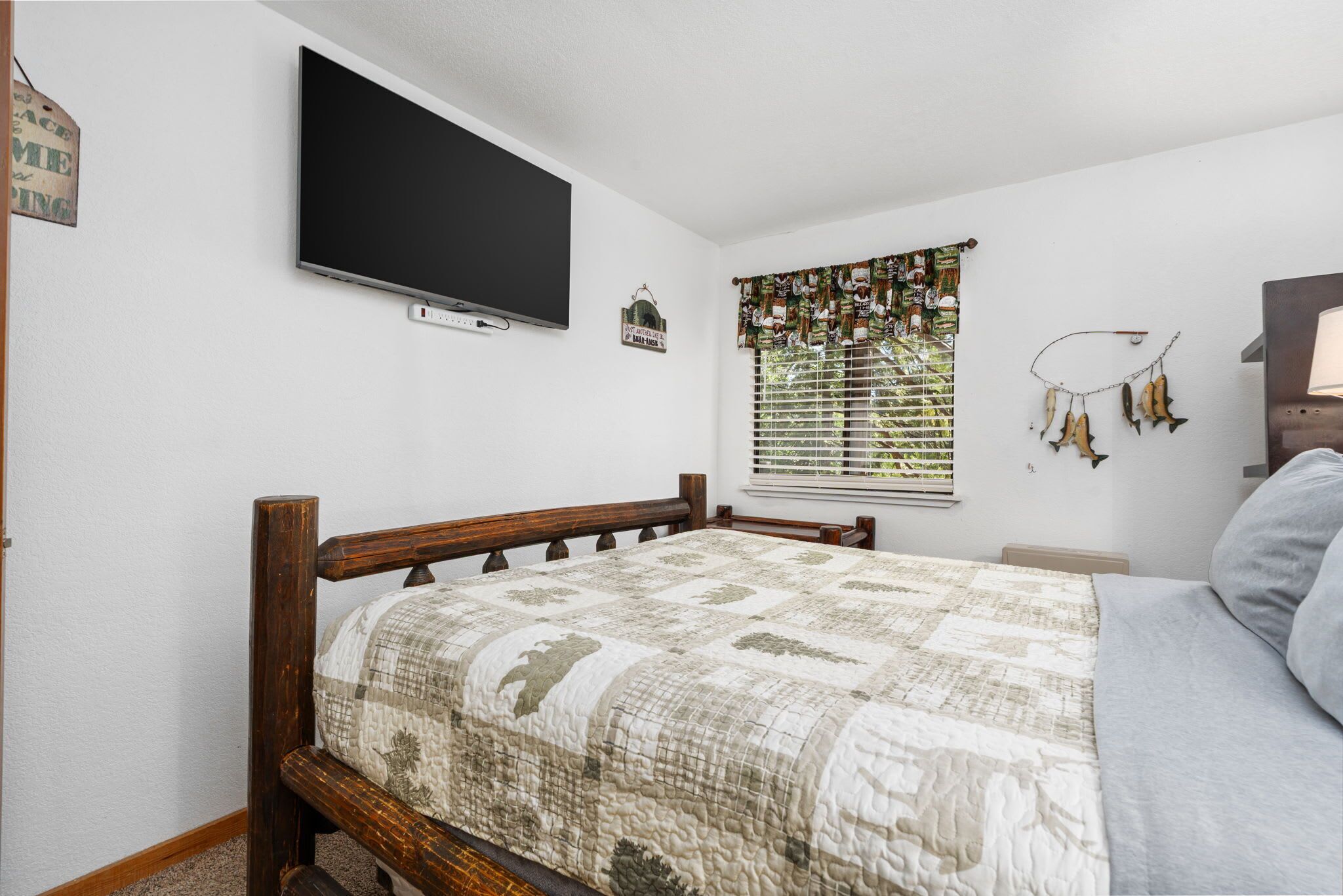 A bedroom with a wooden bed, a flat-screen TV mounted on the wall, a window with patterned curtains, and a wall-mounted dreamcatcher.