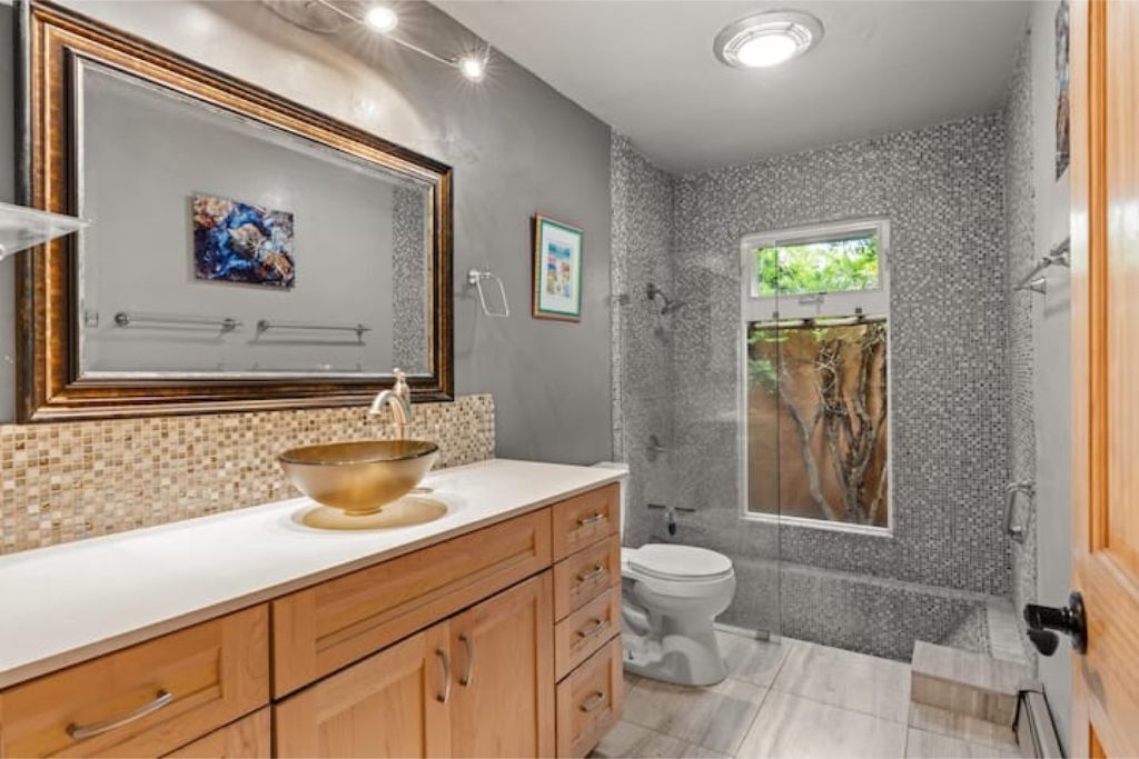 Une salle de bain avec un grand miroir, un lavabo avec un récipient doré, des toilettes et un espace douche avec une fenêtre.