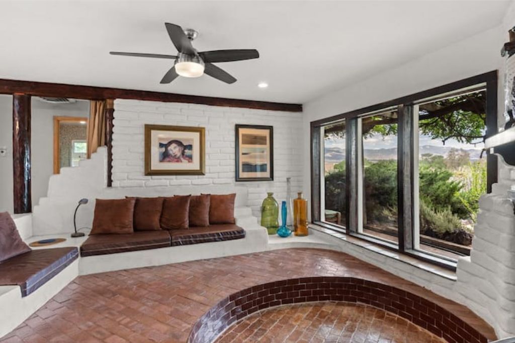 A modern living room with a brick fireplace, a ceiling fan, and large windows offering a view of greenery.