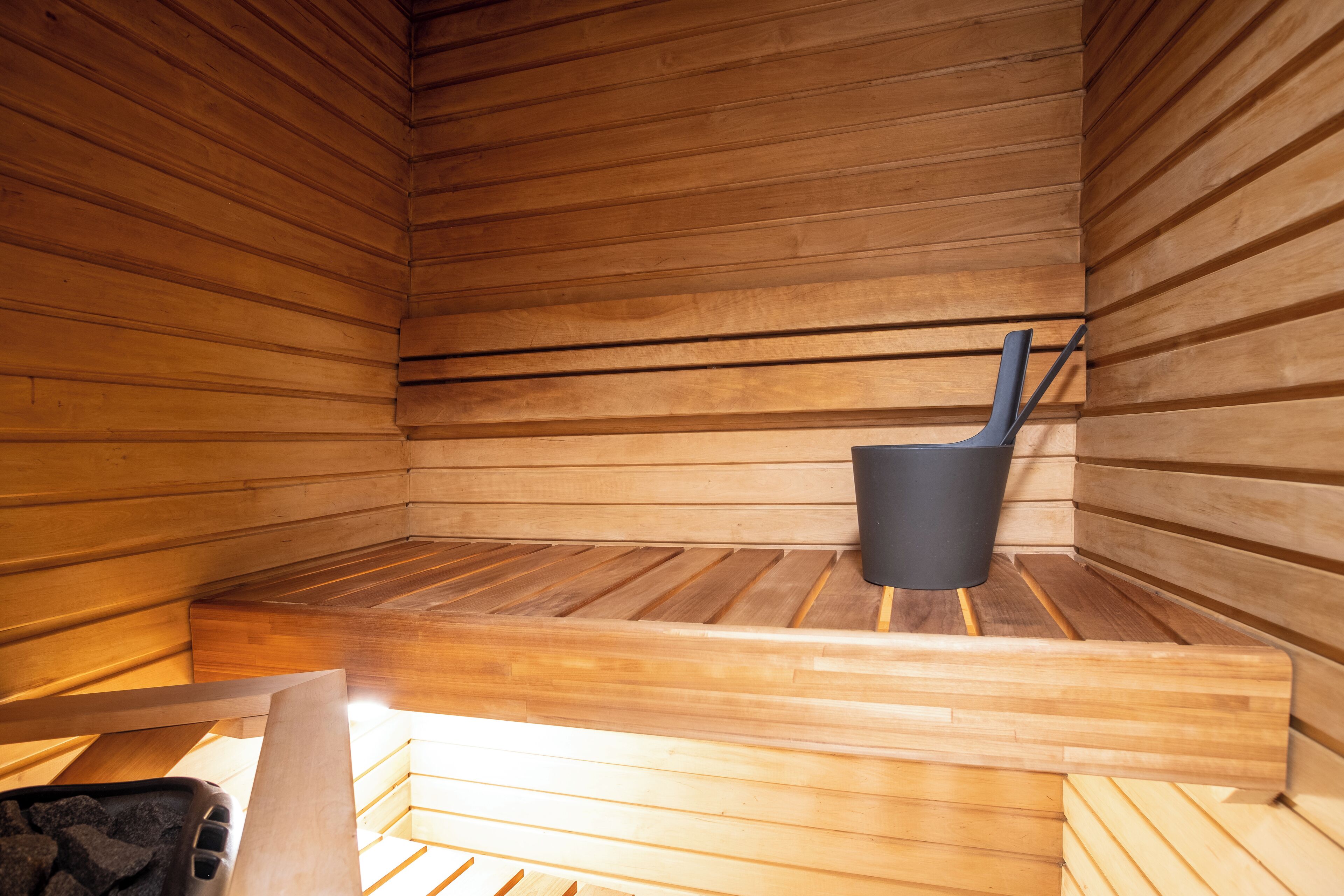 Una sauna con paredes y bancos de madera, un cubo negro y un banco de madera en primer plano.