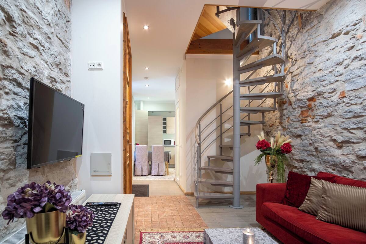 Ein modernes Wohnzimmer mit einem roten Sofa, einem an einer strukturierten Wand montierten Fernseher, einer Wendeltreppe und Blick in den Küchenbereich.