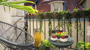Ein Balkon mit einem Korbstuhl, einem Glastisch mit Früchten und Desserts und Blick auf ein Gebäude mit rotem Ziegeldach.