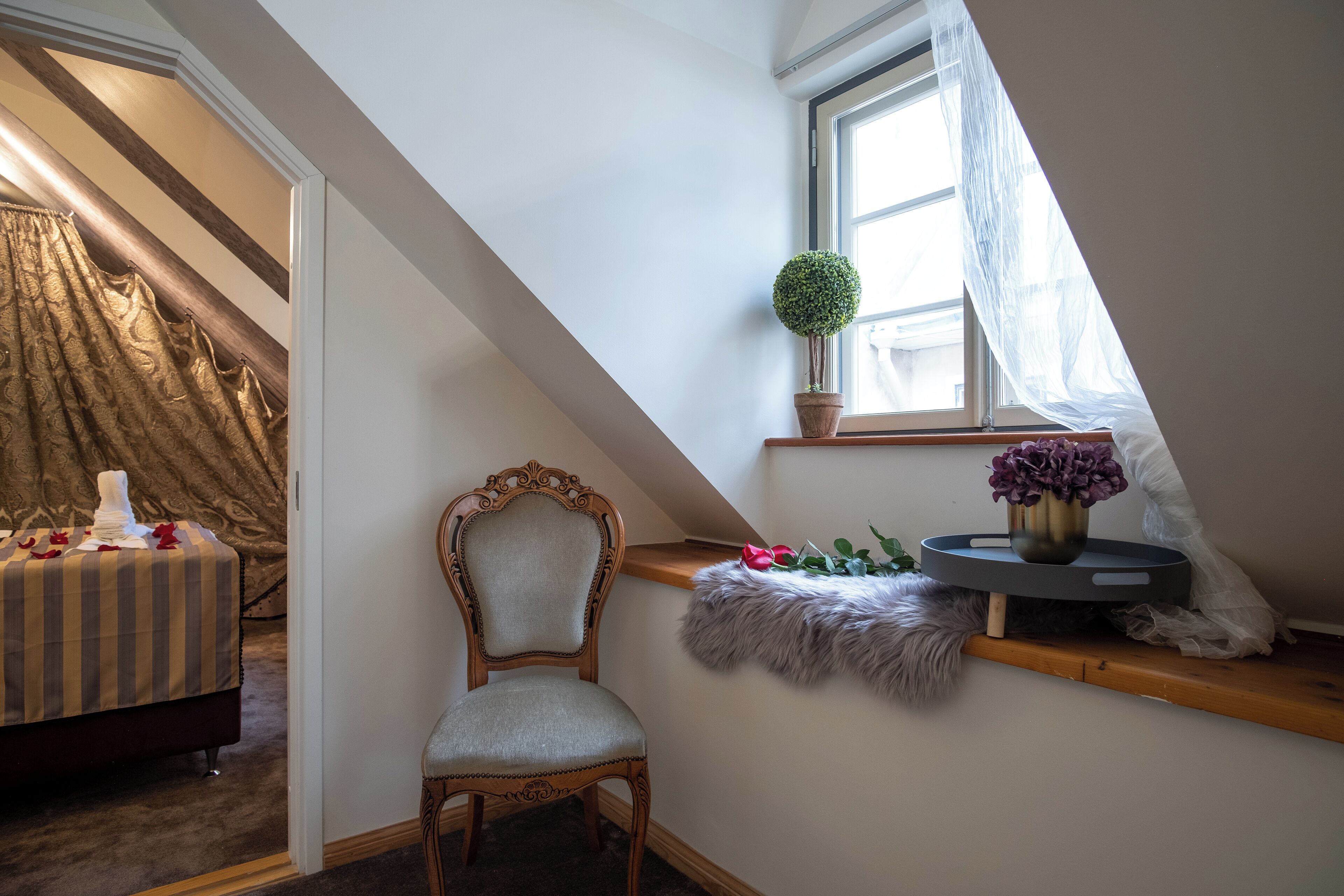 A bedroom with a sloped ceiling, a bed with a striped bedspread, a chair, a window with sheer curtains, and a small shelf with a plant and flowers.