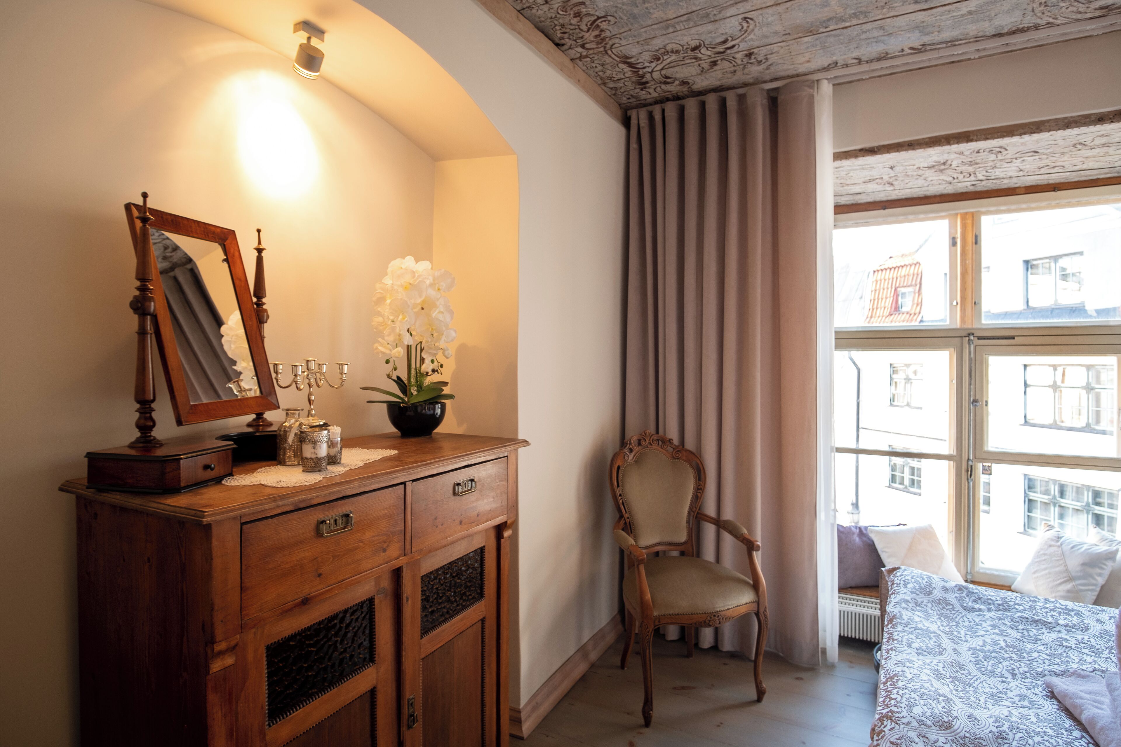 Une chambre à coucher avec une commode en bois, une chaise, un lit et une fenêtre avec des rideaux.