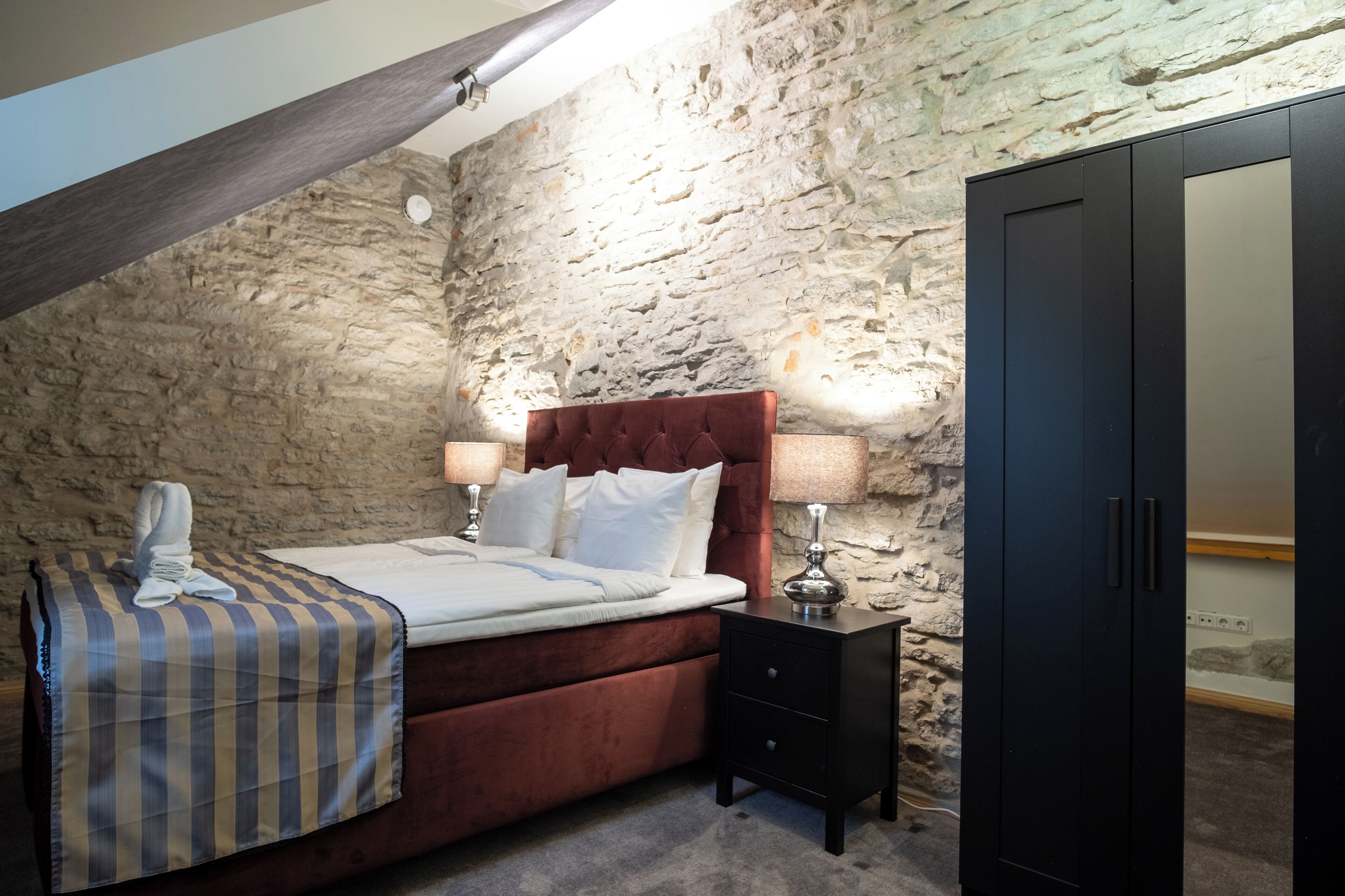 Een slaapkamer met een stenen muur, een bed met een gestreept dekbed, twee bedlampjes en een kledingkast.