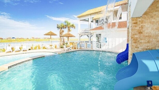 A swimming pool with a water slide, palm trees, and a view of the beach. - A Wave From it All (Corpus Christi)