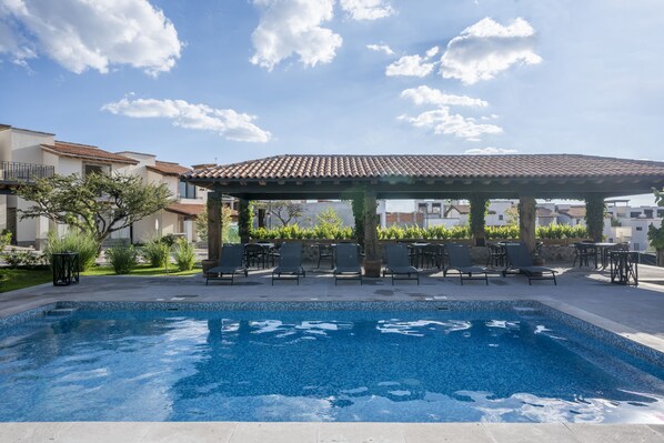 A swimming pool with lounge chairs and a pergola, surrounded by greenery and buildings.