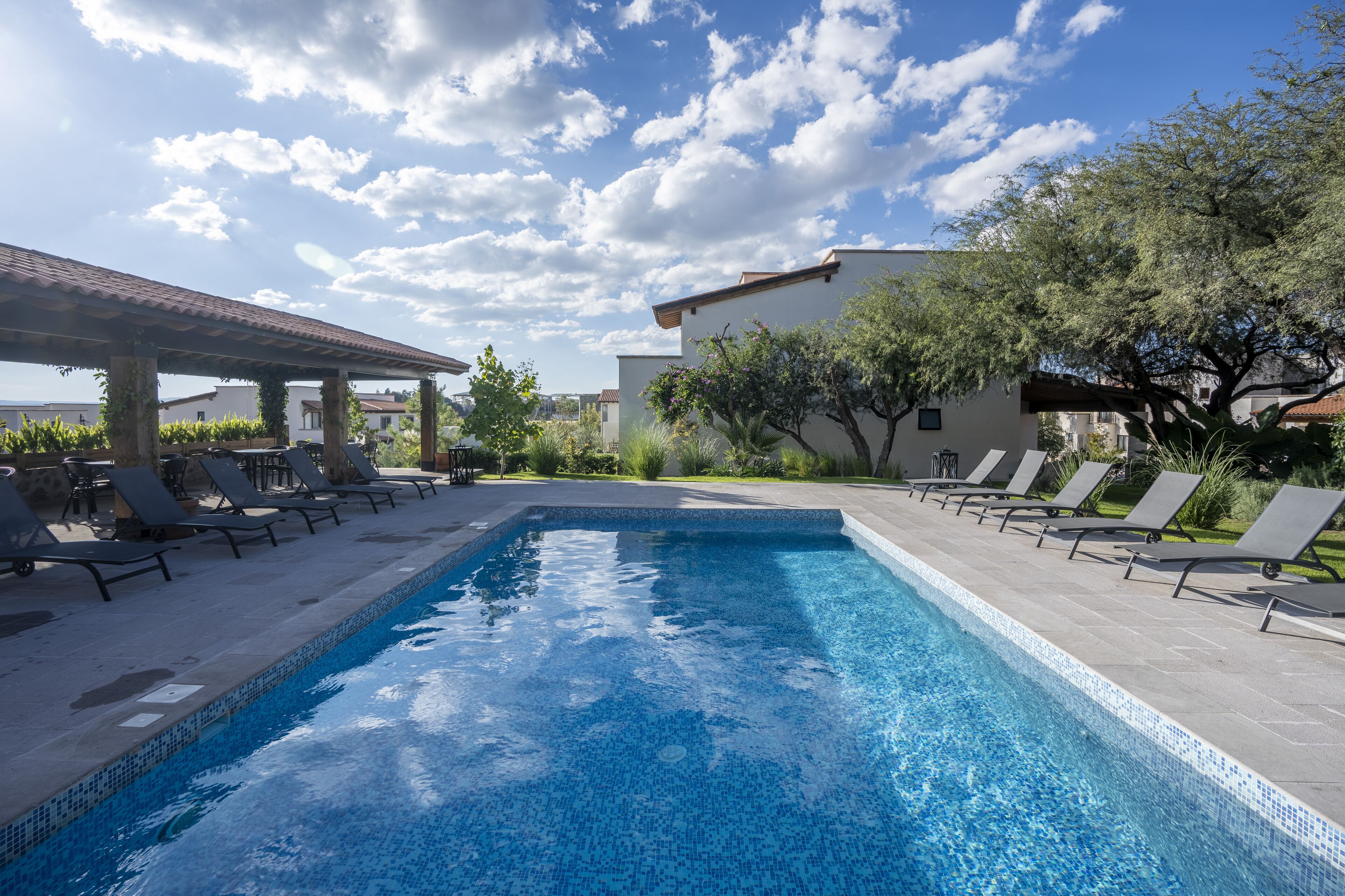 A swimming pool with lounge chairs and a pergola, surrounded by greenery and buildings.