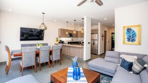 A modern living room with a grey sofa, a wooden coffee table, and a dining area with grey chairs and a wooden table. A kitchen with a fridge, microwave, and sink is visible in the background. - 4BR Private Sanctuary with Pools and Gym (San Miguel de Allende)