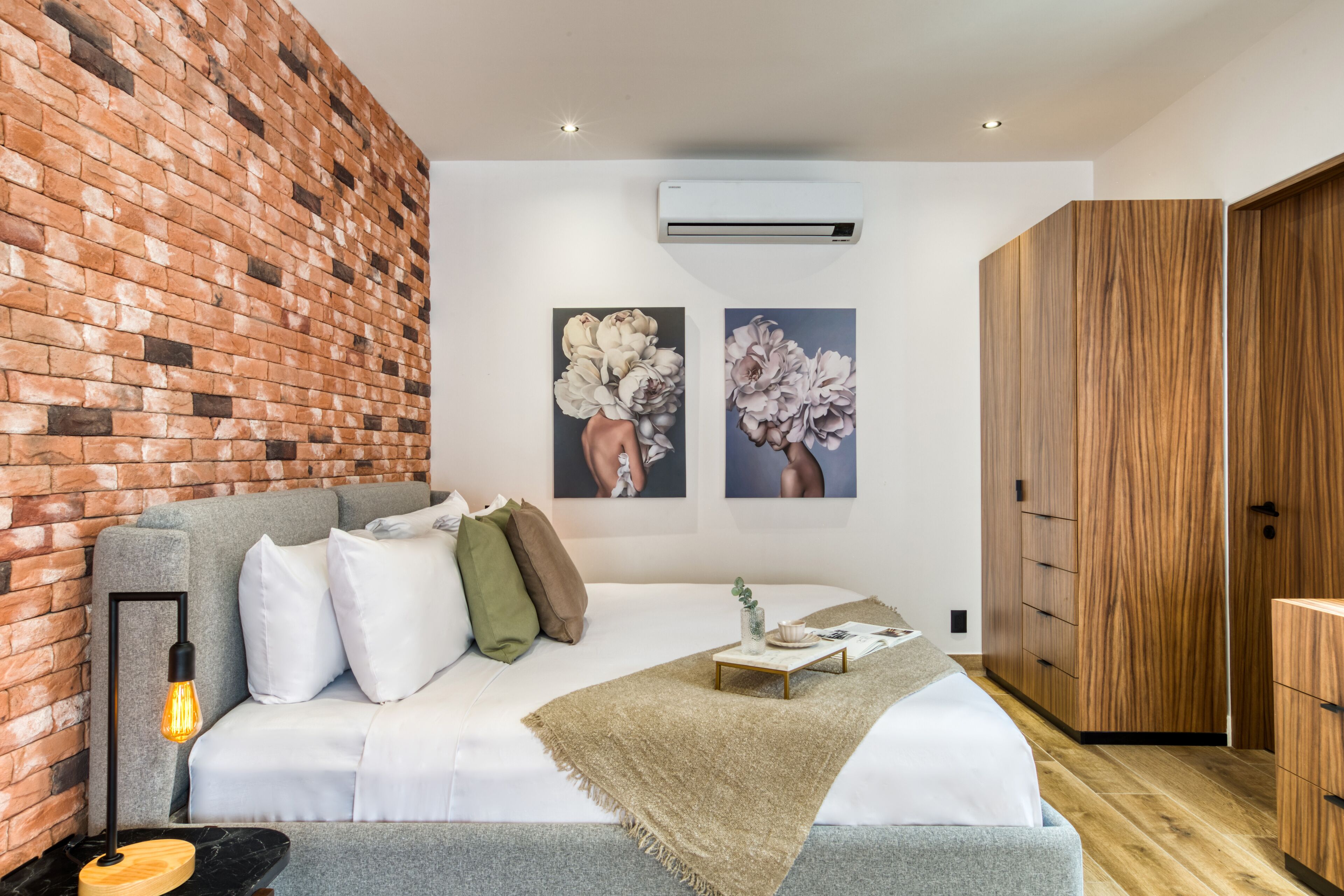 A modern bedroom with a brick wall, a grey sofa, a bed with white bedding, two wall-mounted air conditioning units, and wooden wardrobe.