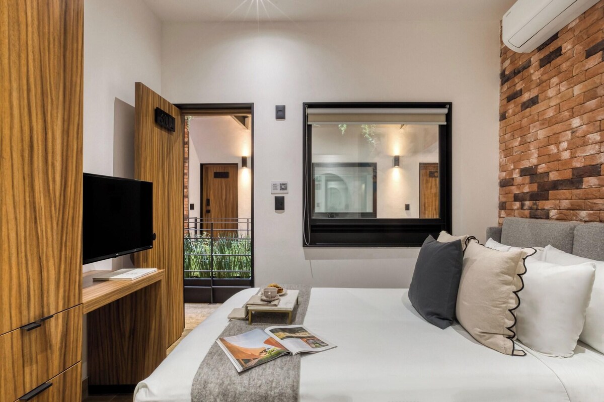 Una habitación de hotel moderna con televisión de pantalla plana, un armario de madera, una cama con ropa blanca y una pared de ladrillo.