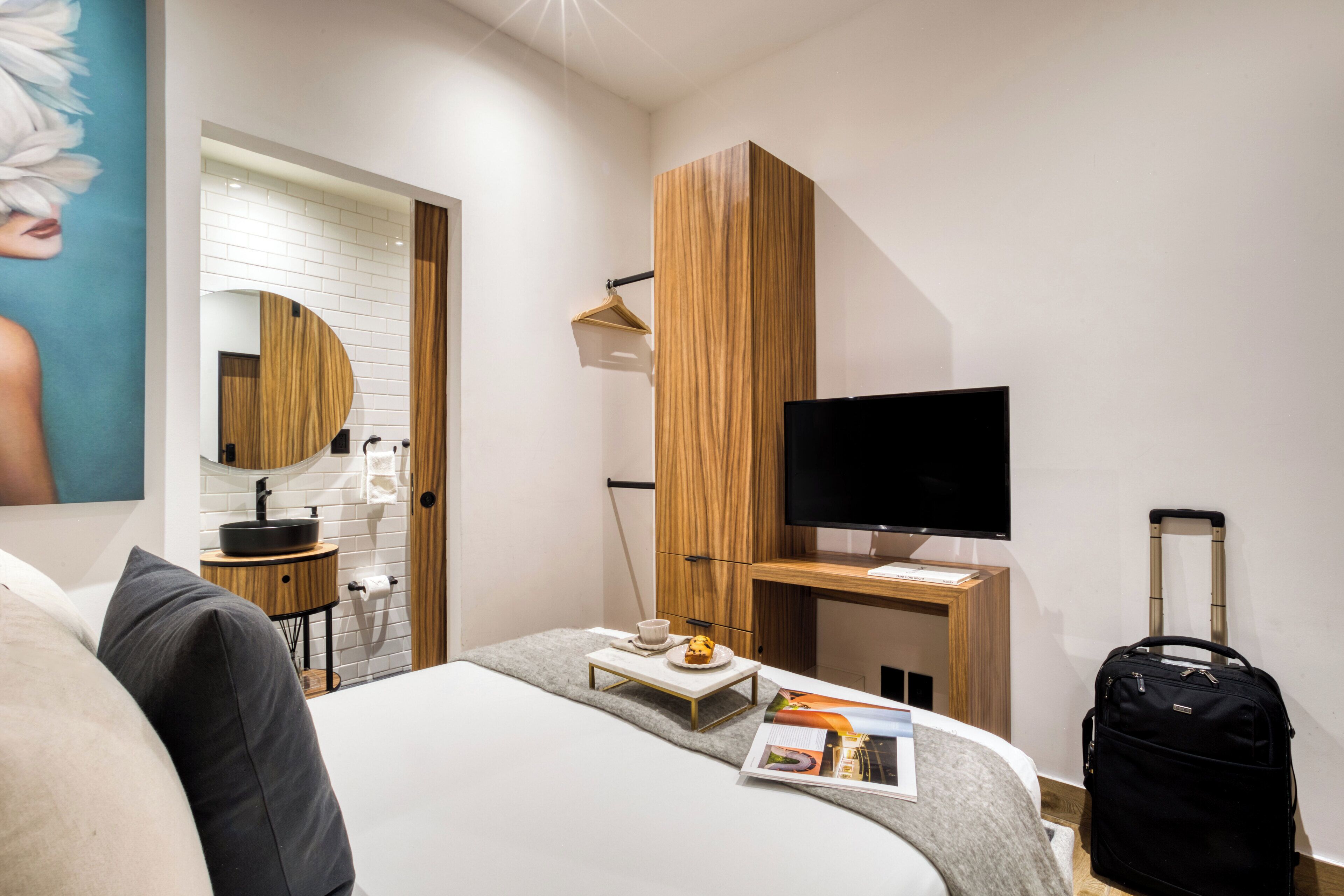 A modern hotel room with a bed, a TV, a small table with a cup and a plate, a suitcase, and a wooden cabinet.