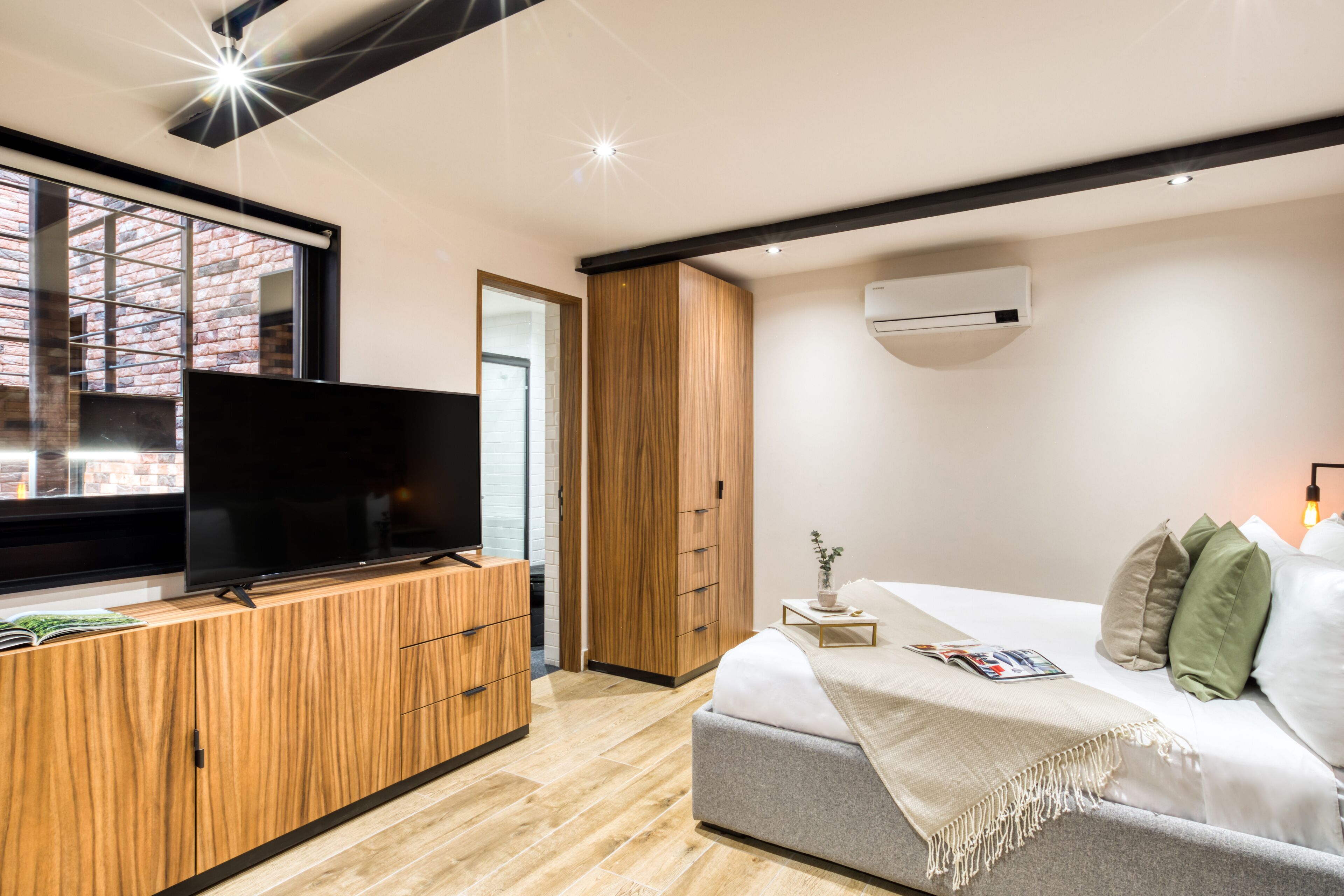 Une chambre moderne avec un téléviseur posé sur un meuble en bois, un lit avec des oreillers et une fenêtre donnant sur un bâtiment en briques.