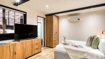 Een moderne slaapkamer met een televisie op een houten kast, een bed met kussens, en een raam met uitzicht op een gebouw van baksteen.
