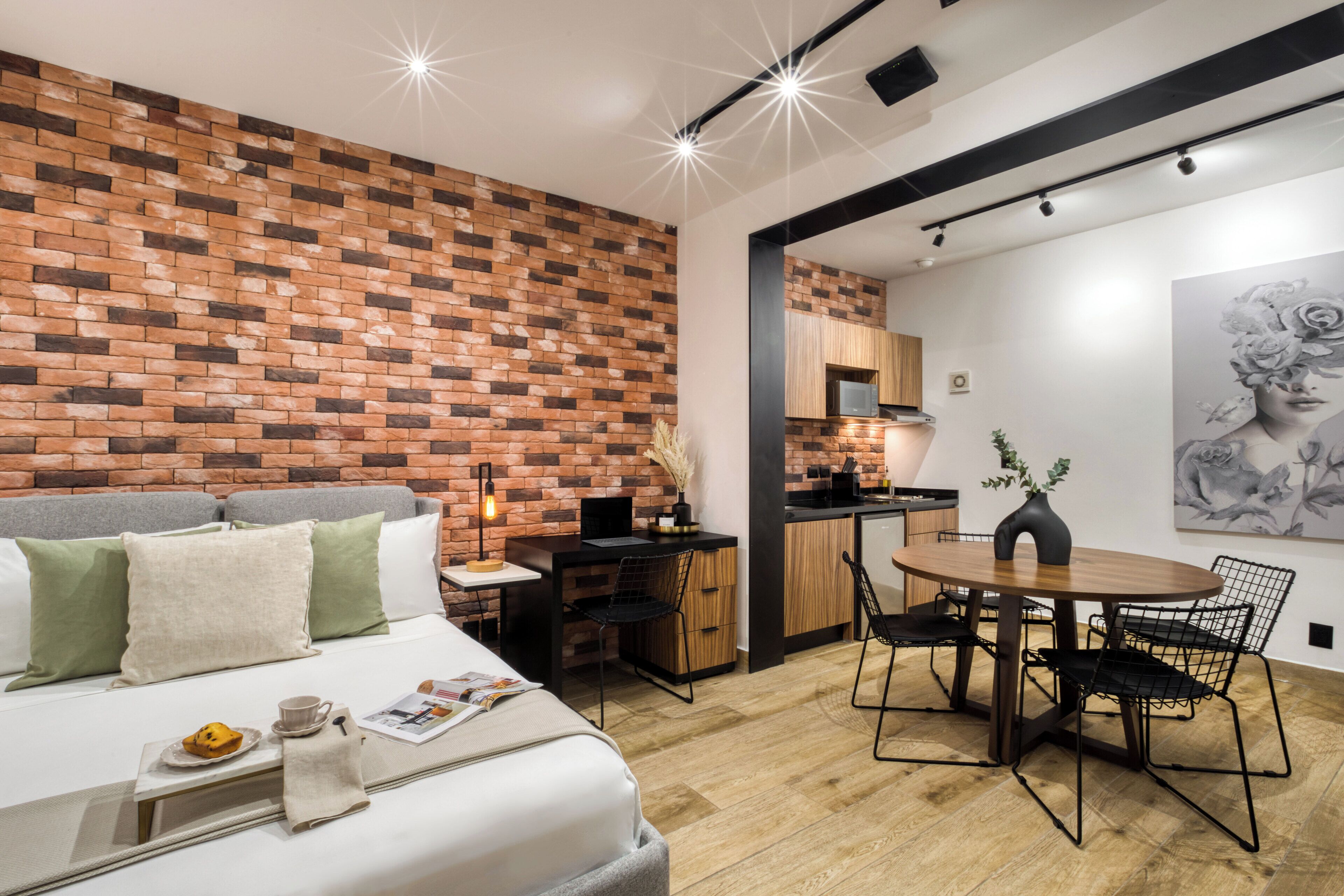 Une chambre d’hôtel moderne avec un mur en briques, un lit agrémenté d’oreillers verts et blancs, une table à manger avec des chaises, et un coin cuisine visible à travers une porte ouverte.