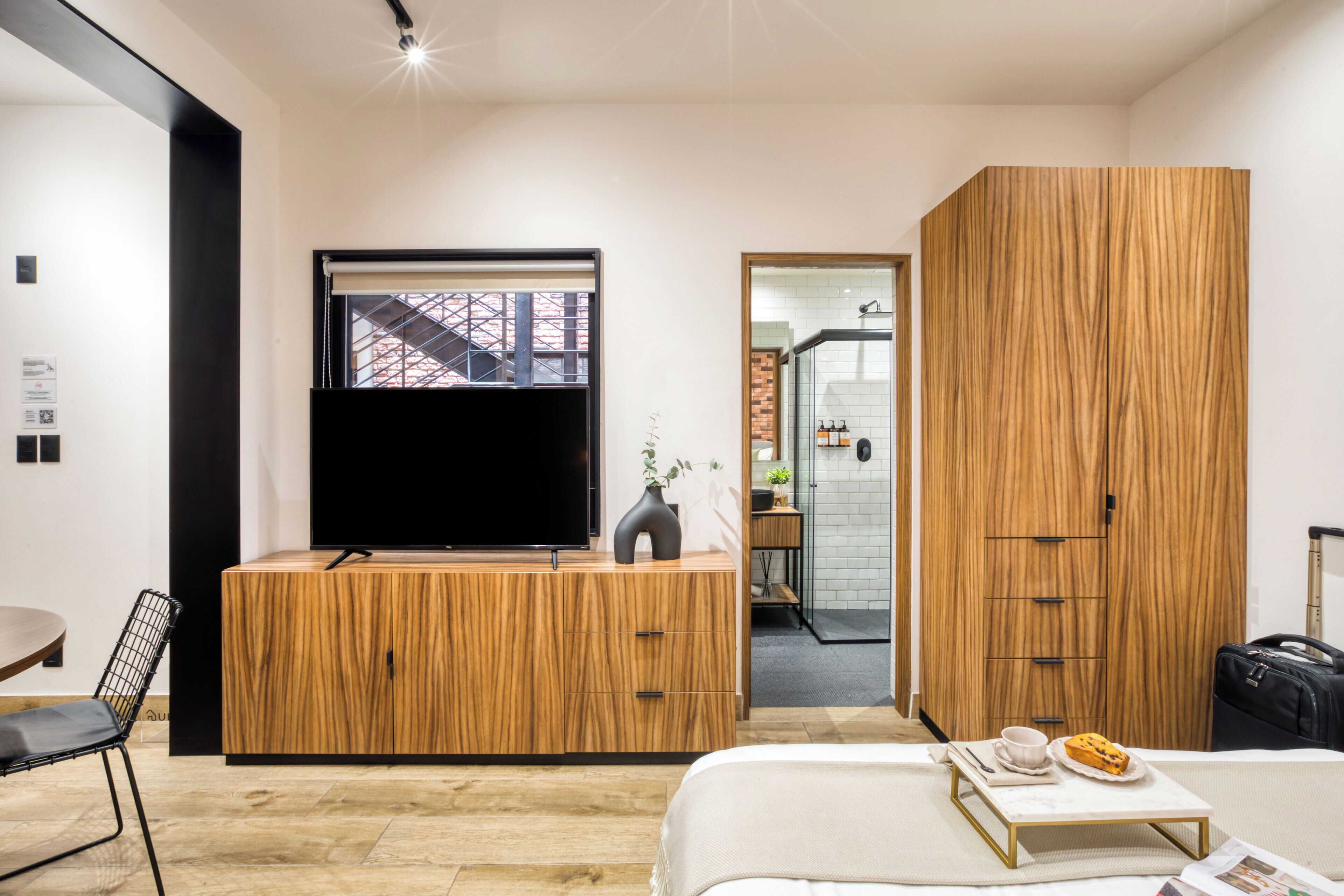 A modern hotel room with a wooden cabinet, a flat-screen TV, a bed with a tray table, and a suitcase.