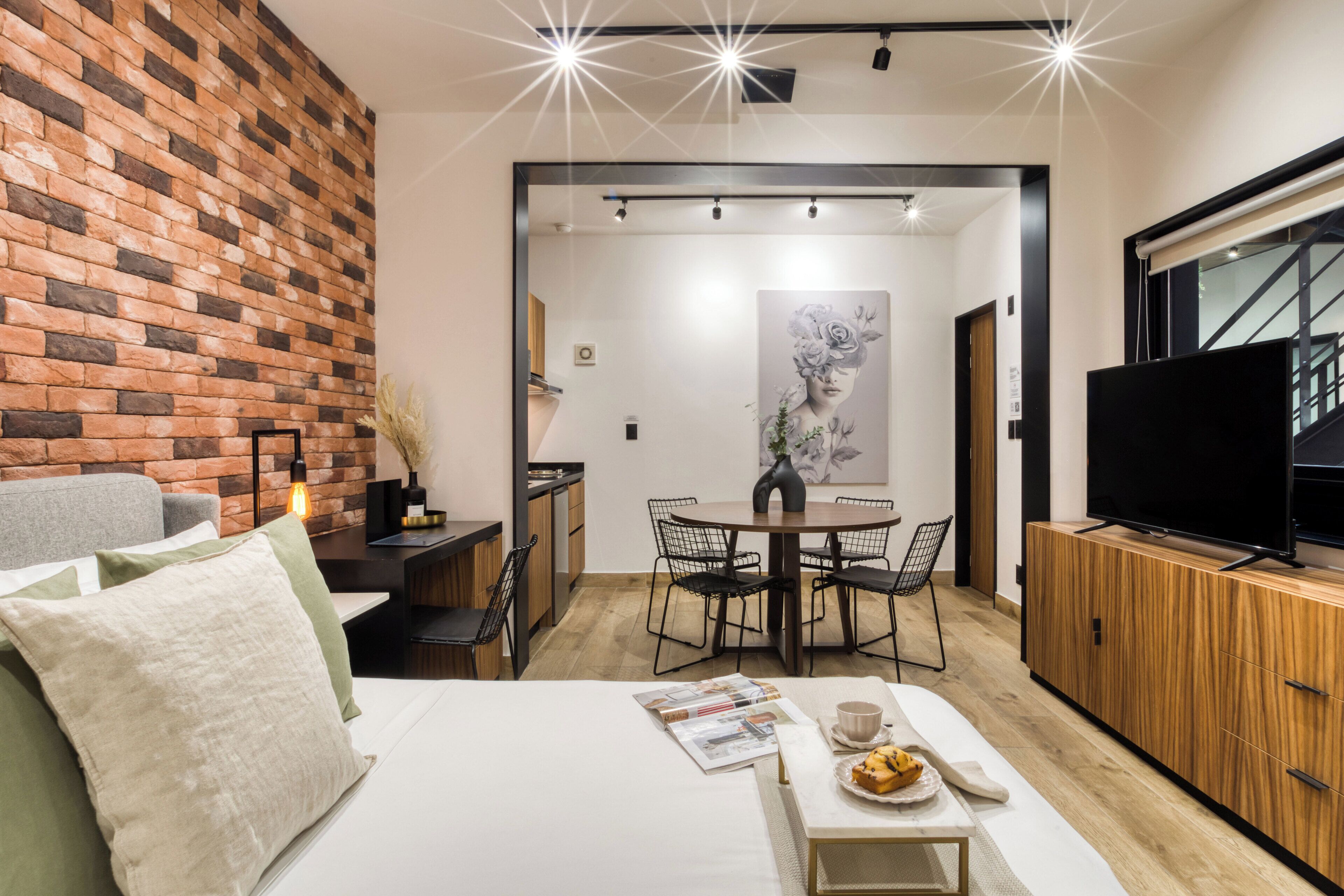 Habitació d'hotel moderna amb paret d'accent de maó, zona de menjador amb taula rodona i cadires, televisió de pantalla plana i llit amb capçalera blanca.