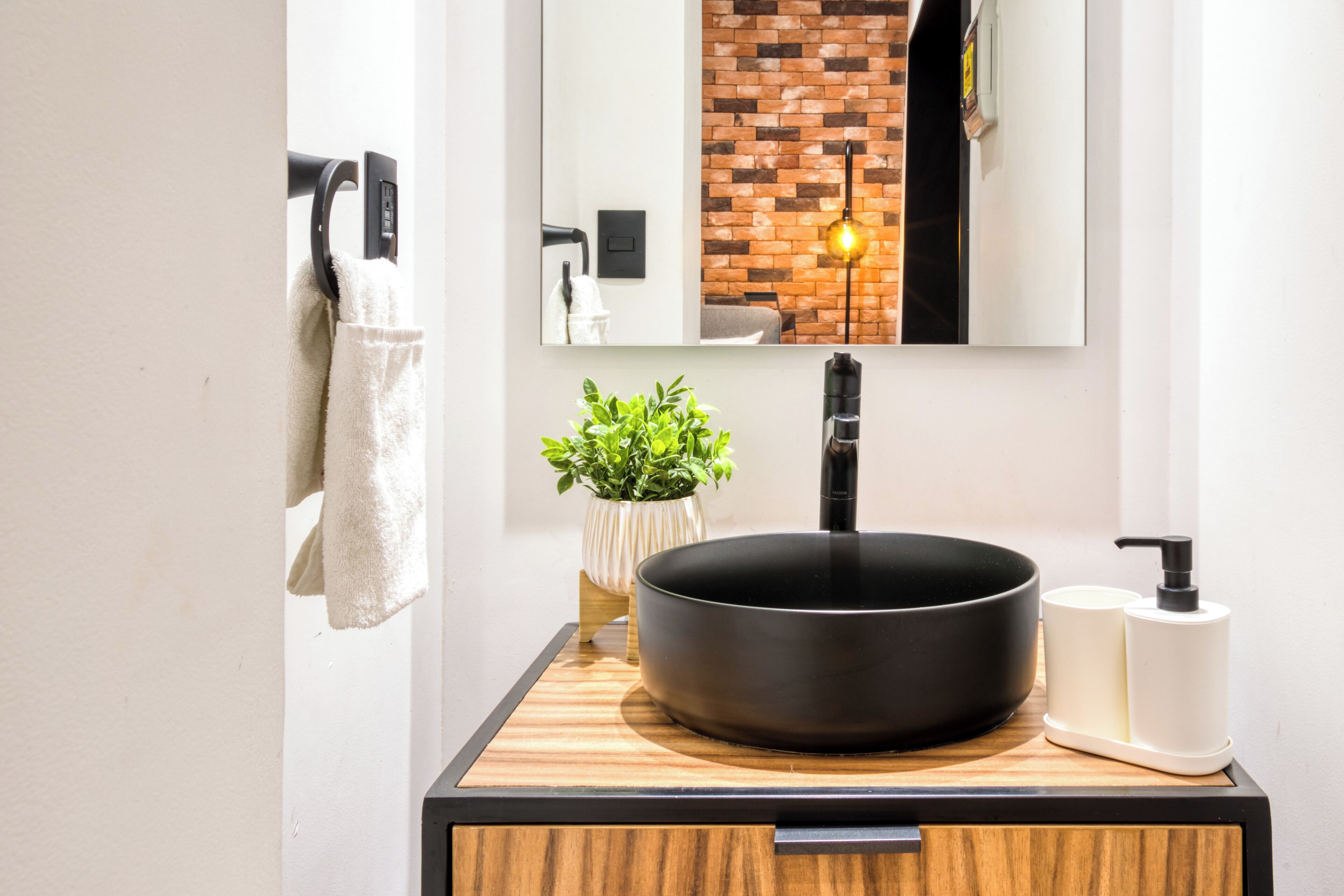 A modern bathroom with a black sink, wooden vanity, a potted plant, a towel rack, and a mirror reflecting a brick wall.