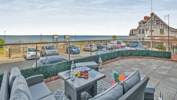 Eine Strandterrasse mit Sitzgelegenheiten im Freien, Blick auf das Meer, geparkte Autos und ein Gebäude im Tudor-Stil.