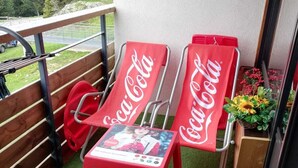Un balcón con dos sillas rojas con la marca Coca-Cola, una mesita con un cuadro y vistas a árboles y una carretera.