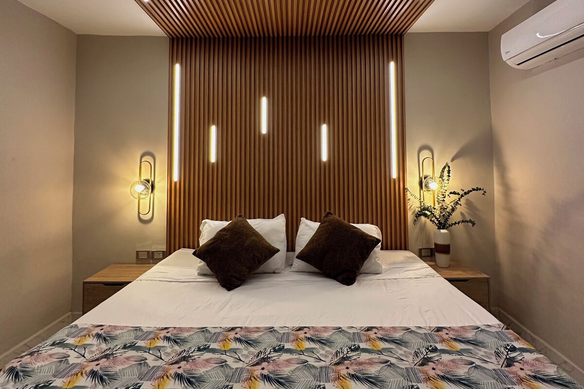 Quarto moderno com teto de ripas de madeira, cama grande com roupa de cama estampada, mesas de cabeceira com luminárias e unidade de ar condicionado.