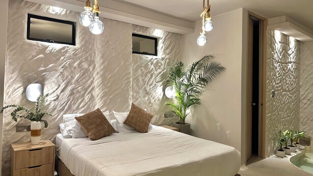 A modern bedroom with a large bed, bedside tables, a mirror, and a wall-mounted light fixture. - HOTEL  BALI AT POBLADO (Medellín)