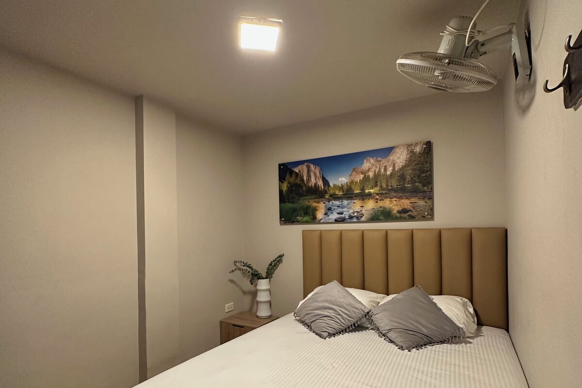 Quarto com cama, mesa de cabeceira, luminária na parede, um quadro e luz de teto.