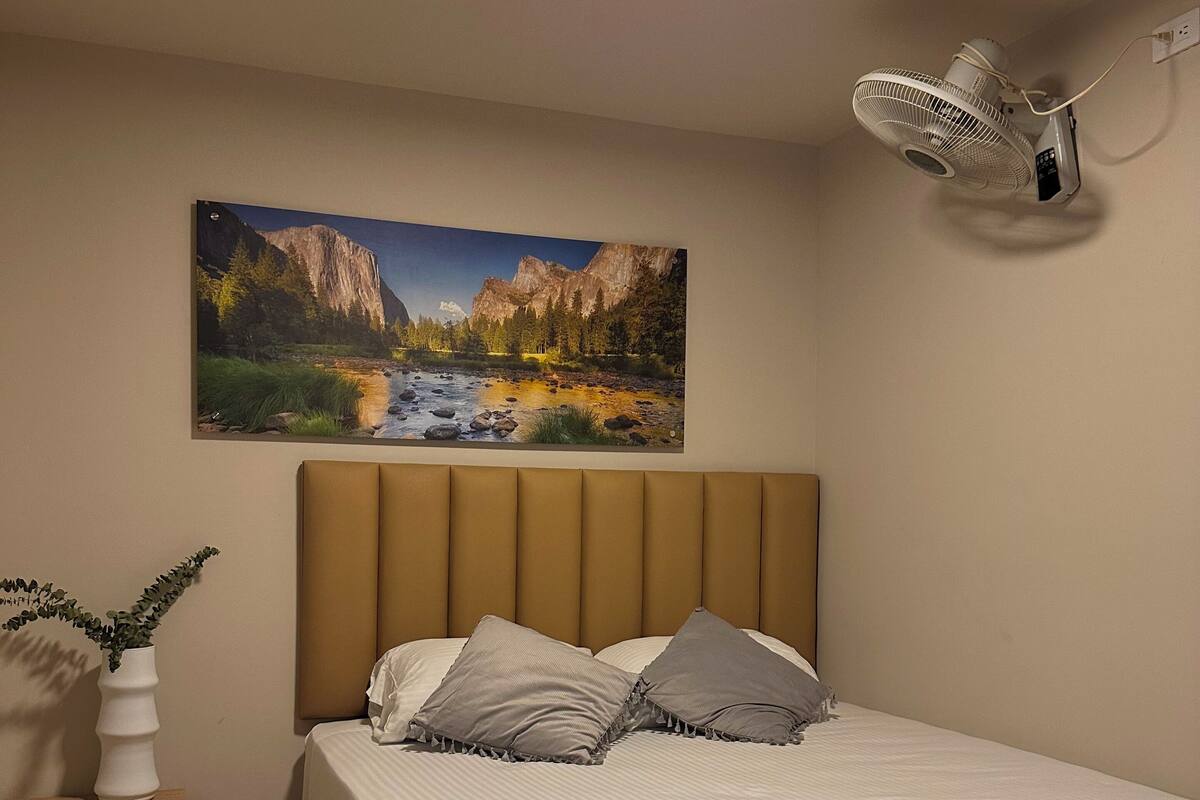 A bedroom with a bed, a nightstand, a wall-mounted fan, and a painting of a landscape.