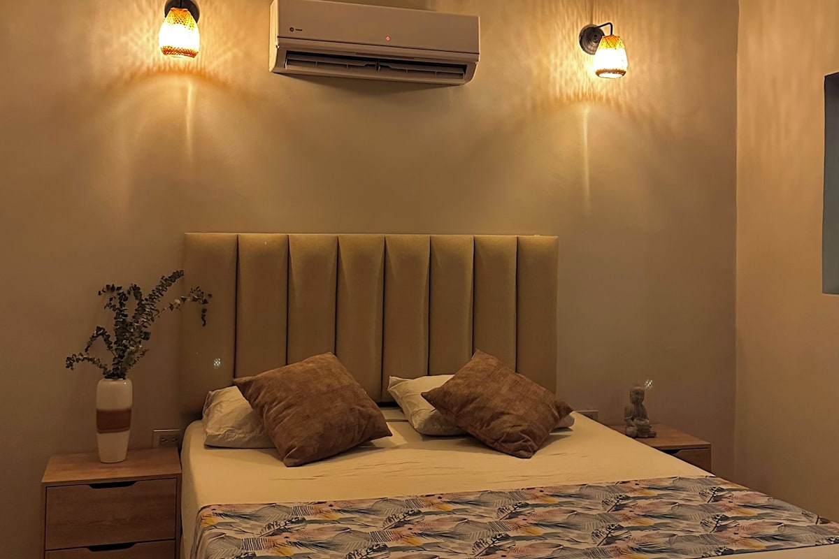 A bedroom with a bed, two wall-mounted lamps, an air conditioner, a nightstand with a vase, and a wooden drawer.