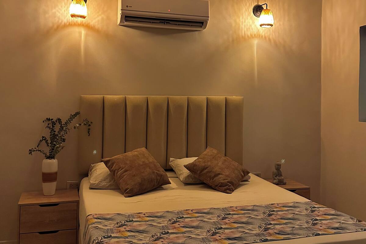 A bedroom with a bed, two wall-mounted lamps, an air conditioner, a nightstand with a vase, and a wooden drawer.