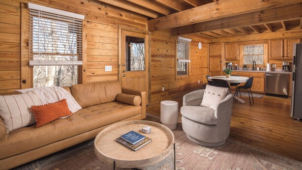 A cozy cabin interior with a wooden interior, a sofa, a round coffee table, a chair, and a kitchen area.
