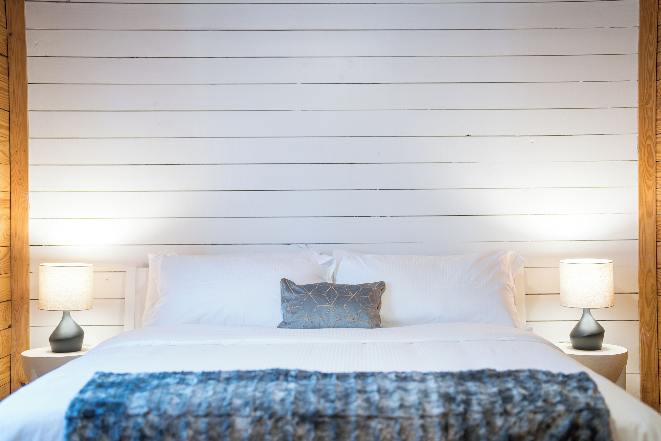 A neatly made bed with white linens, a blue patterned throw, and a geometric pillow. There are two bedside lamps on wooden nightstands.