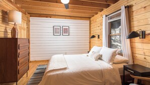 A wooden cabin bedroom with a bed, dresser, lamp, and framed pictures on the wall.