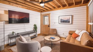 A cozy room with a wooden ceiling, a mounted TV, a sofa, a chair, and a small table with a book and a plant.
