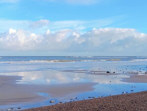 A beach with pebbles and wet sand, reflecting the sky and clouds.