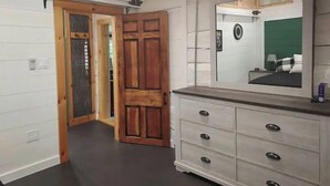 Una habitación con puerta de madera, cómoda con cajones y espejo que refleja una cama con cabecera verde.