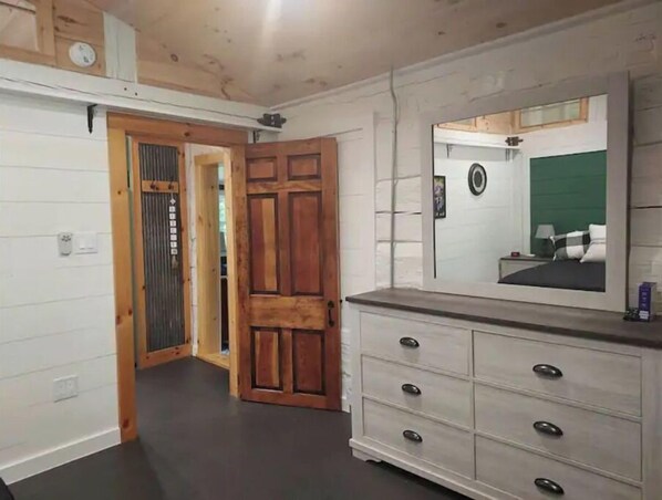 A room with a wooden door, a dresser with drawers, and a mirror reflecting a bed with a green headboard. - Little Cabin in the Woods (Morganton)