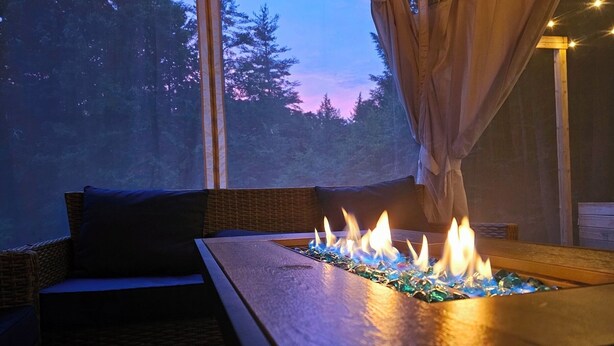 A cozy seating area with a fire feature in the center, a window with curtains, and a view of trees. - Spacious White Mt. Cabin - Outdoor Patio & Fireplace (Thornton)