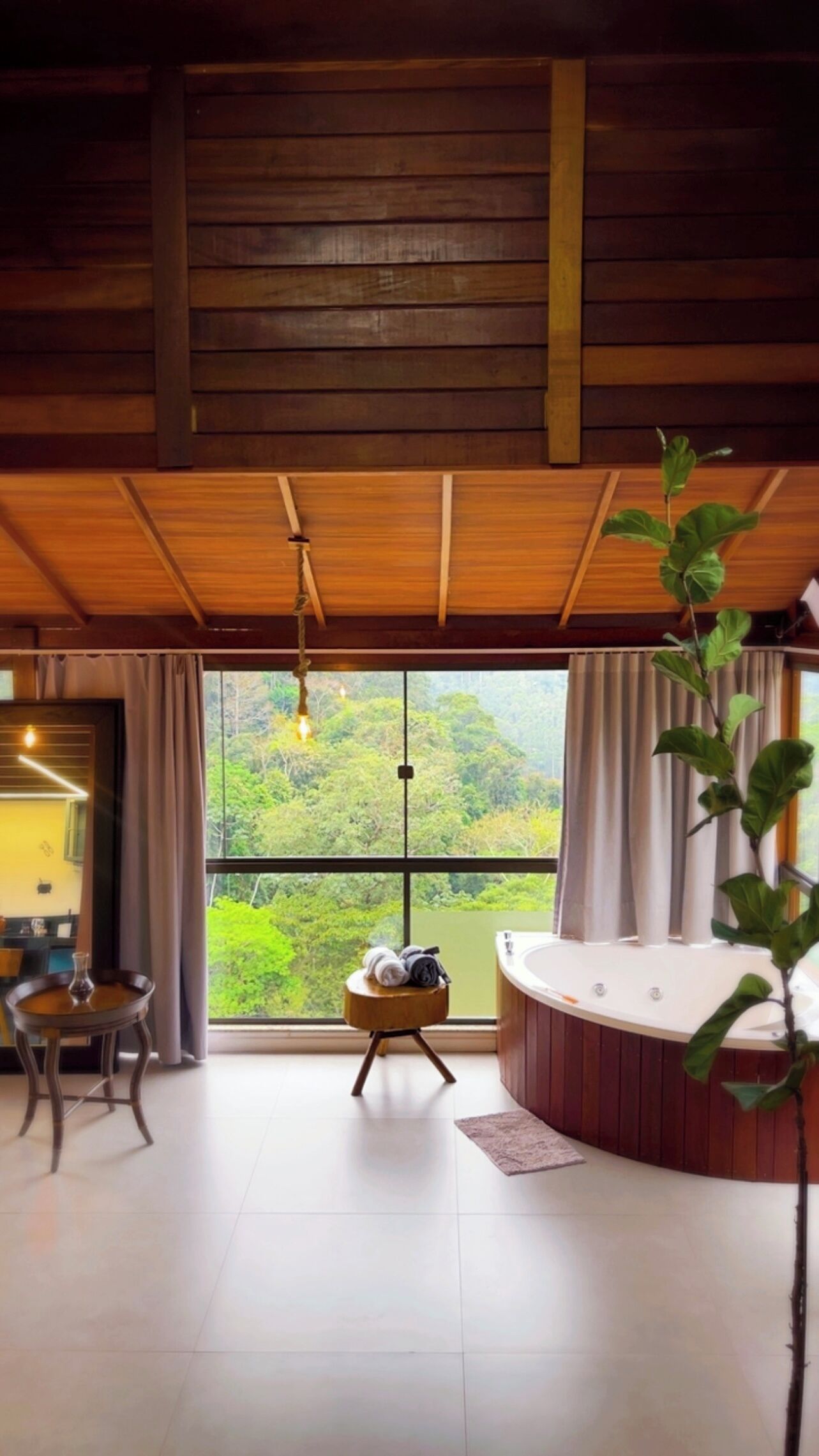 Une pièce avec un plafond en bois, une grande fenêtre avec des rideaux, un jacuzzi rond, un petit tabouret en bois avec des serviettes et une table sur laquelle est posé un vase.