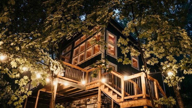 A multi-level wooden structure with a stone base, surrounded by trees and illuminated by string lights.