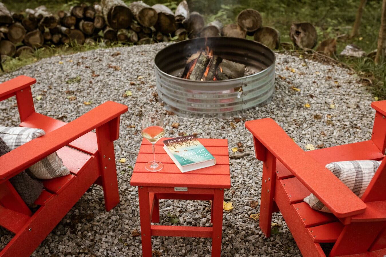 A fire pit surrounded by red Adirondack chairs and a small table with a book and glass of wine.