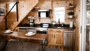Une cuisine compacte avec des meubles en bois, un four à micro-ondes, une cuisinière et un évier. On y trouve une table à manger avec des chaises, ainsi qu’un escalier menant à un autre niveau.