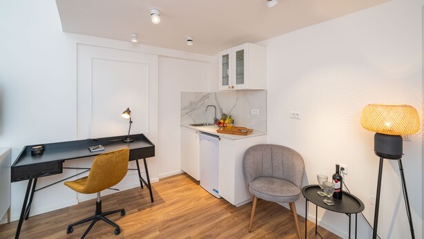 A compact living space with a wooden floor, a desk with a chair, a small kitchen area with a sink and cabinets, and a floor lamp. - Villa Aura - Executive Business room (Dubrovnik)