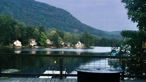 A deck with a glass table and a glass of red wine, overlooking a lake and mountains.