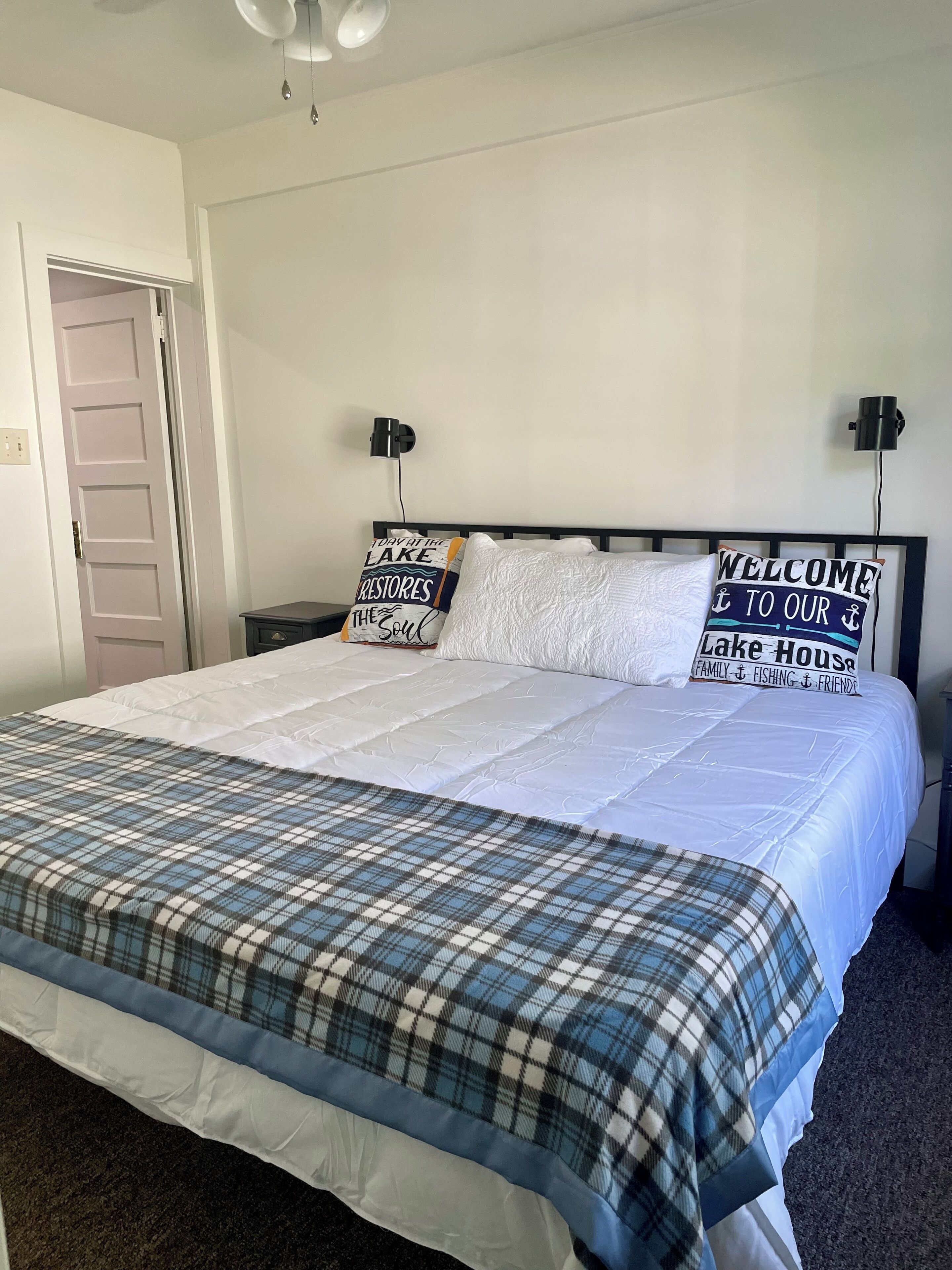 A neatly made bed with a plaid bedspread, two pillows with text, a nightstand with a lamp, and a door in the background.