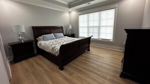Une chambre à coucher avec un lit en bois foncé, deux tables de chevet avec des lampes, un ventilateur de plafond et une grande fenêtre avec des stores.