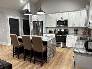 A modern kitchen with white cabinets, stainless steel appliances, and a granite island with bar stools. - Modern 3 BR home, interior lot near Lake Keowee, Clemson, Walhalla, Seneca. (Seneca)