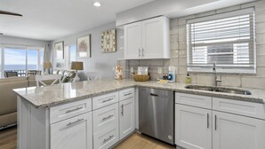 A modern kitchen with white cabinetry, granite countertops, a stainless steel dishwasher, and a window with blinds. - NEW! 2/2, IRB-Beach/Pool View, Sleeps 6, unit F (Indian Rocks Beach)