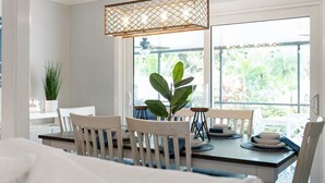 A dining area with a table set for four, surrounded by white chairs, a hanging light fixture, and a view of greenery outside. - Coastal Retreat w/ Porch, Games, Pet Friendly! (Naples)