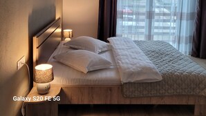 Una cama bien hecha con sábanas blancas y un edredón acolchado, una lámpara de noche y una composición de fotos en la pared.